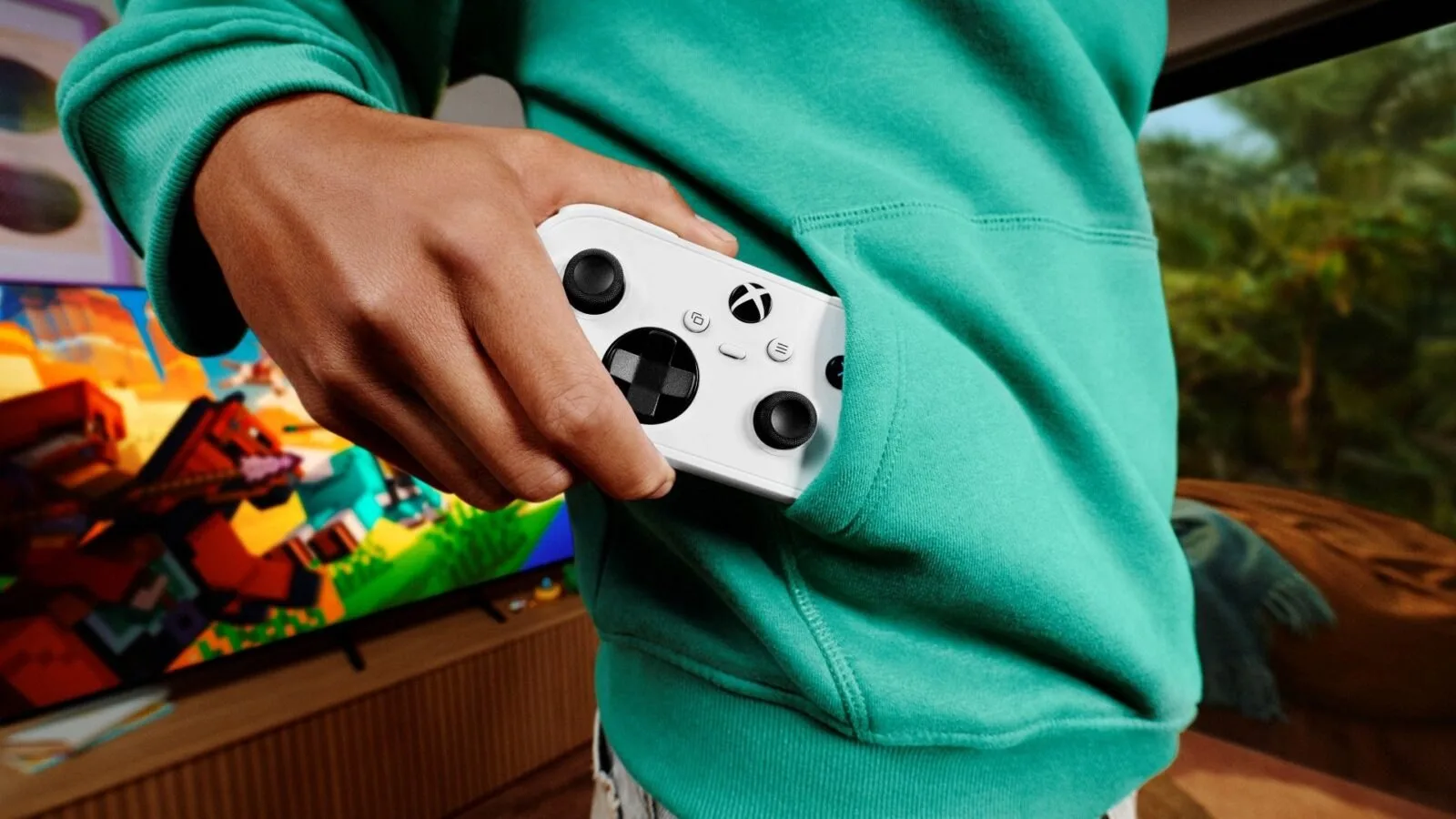 Beyaz Xbox Nano Controller bir kişinin kapüşonlu sweatshirtünün cebine sığıyor, arkada televizyonda Minecraft görüntüsü